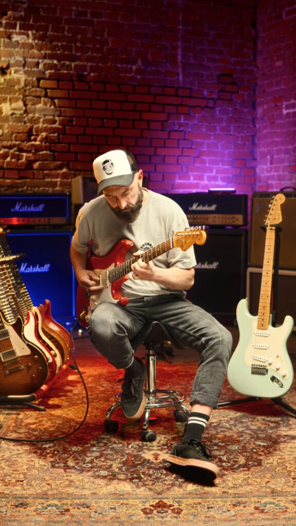 Mężczyzna siedzący na krześle z gitarą, w chwili relaksu, z uśmiechem, oddający się muzyce.