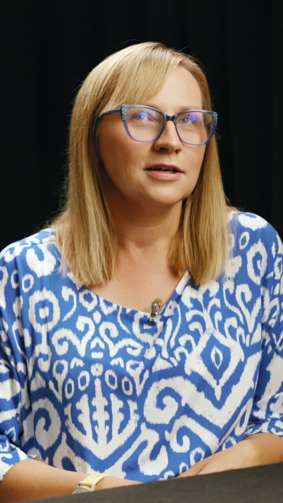 Kobieta w okularach, ubrana w niebiesko-białą koszulę, uśmiecha się do kamery.