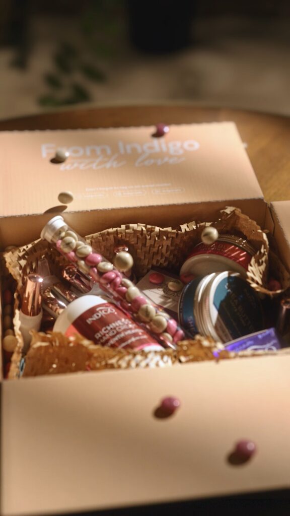 Pudełko z kosmetykami Indigo Nails, w tym balsamy i cukierki M&M's, leży na stole.